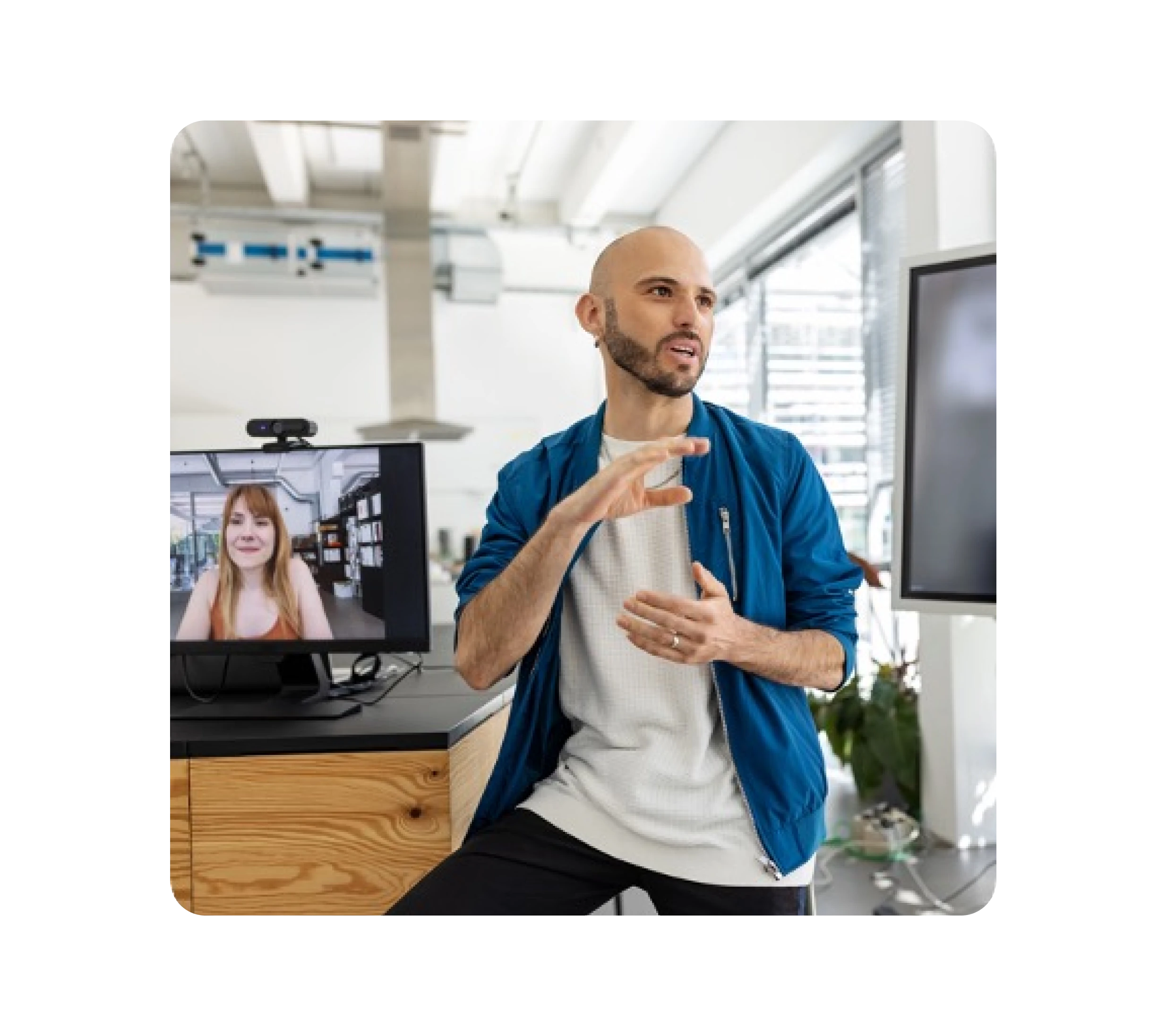 A man gestures as he speaks in an office while a monitor beside him shows a colleague on video